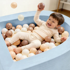 A smiling toddler in beige lies in the Tiny Land® Blue PlayNest Ball Pit by Tiny Land INC, happily tossing 200 ocean balls in brown, cream, and white throughout the light blue play area.|blue