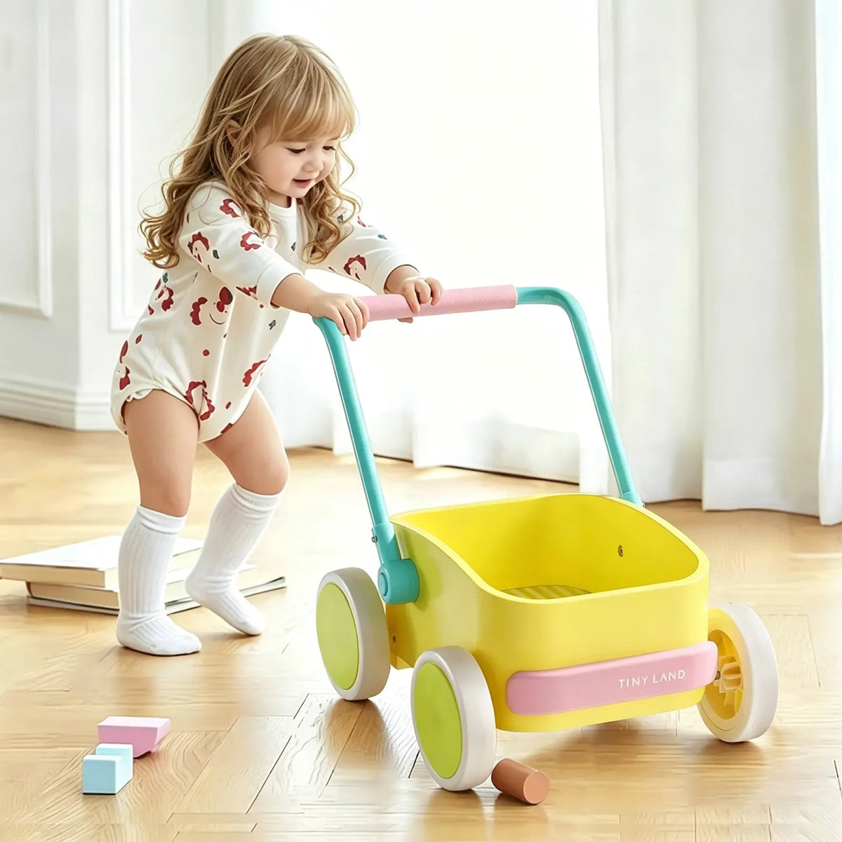 A young child in a patterned onesie and knee-high socks pushes the Test Tiny Land® JoyRide Baby Walker with Blocks - Spring Color across a sunlit wooden floor, surrounded by blocks, books, and a wooden storage box.