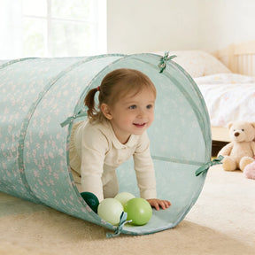 A young child smiles while crawling through the Tiny Land® Adventure Play Tunnel - Green by Tiny Land, with green and white plastic balls inside. A bed and teddy bear can be seen in the background.|green