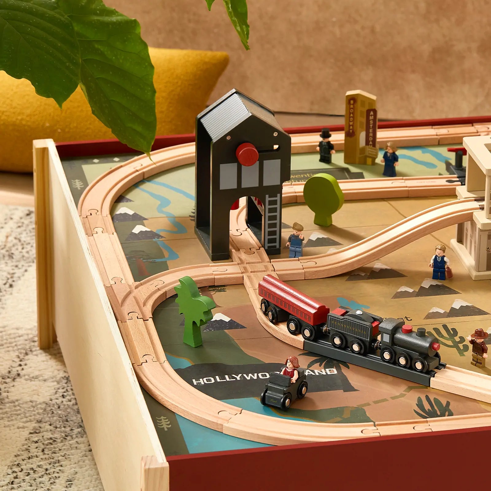 The Tiny Land® 1920 Railway Adventure Train Table by Tiny Land INC features tracks, a black and red vintage locomotive, small figures, buildings, trees, and a Hollywoodland sign on a playset table with plant and yellow cushion accents in the background.