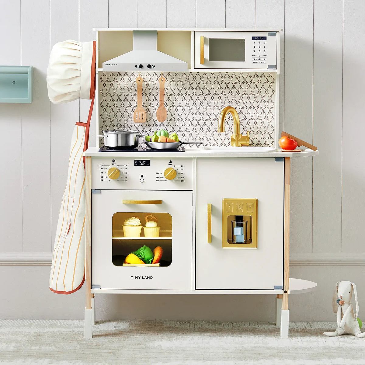 The Tiny Land® PopJoy Play Kitchen by Tiny Land includes a chef hat, oven, stove, sink, utensils, toy food, microwave, and faucet. Set against a light wall with a toy rabbit on the right floor.