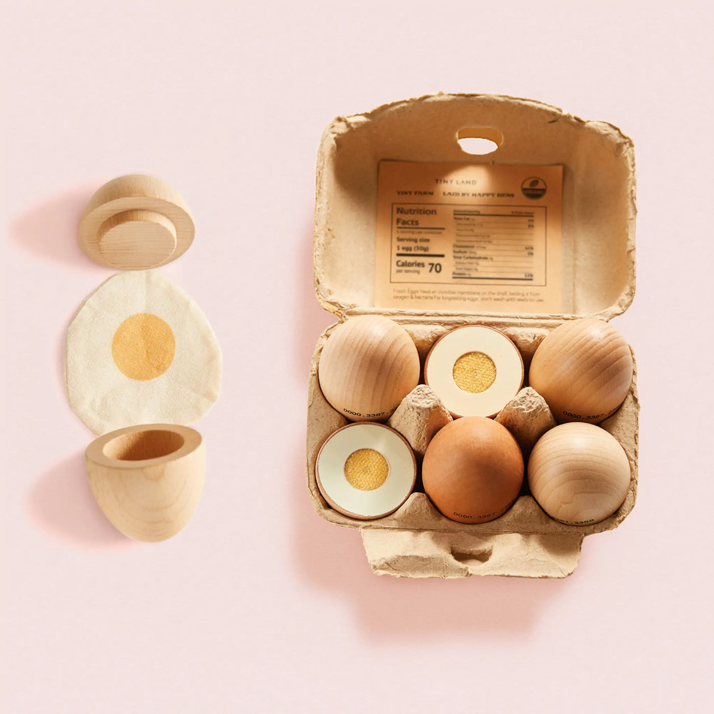 A carton of six wooden eggs from the Tiny Land® Egg Combo Playset by Tiny Land Play Food—some whole, some halved to show yolks—rests on a pink surface beside two opened eggs and a cloth with yellow yolk print, perfect for toddler pretend play.