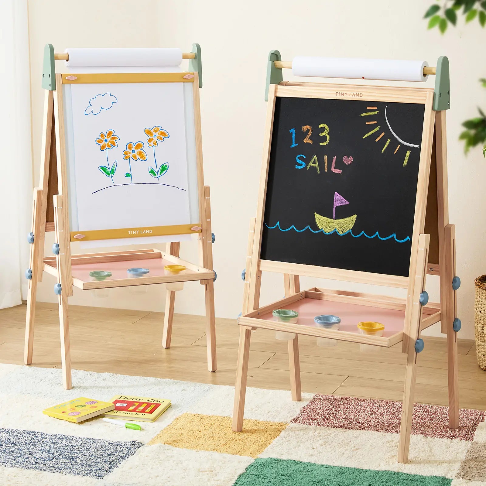 Two Tiny Land® 3-in-1 Art Easels for Kids - Morandi stand on a colorful rug. One shows flower art on paper, the other displays “123 SAIL” and a boat under the sun in chalk. Paint trays are attached below each drawing surface.