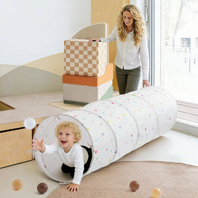A smiling child crawls through the Tiny Land® Adventure Play Tunnel - Colorful Polka Dots as a woman looks on, both enjoying playtime in a bright room filled with soft toys and modern furniture.|white