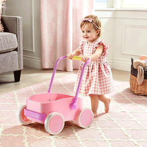 A toddler in a pink gingham dress pushes the Tiny Land® 3-in-1 Wooden Baby Walker Wagon - Pink across a pink rug in a cozy, sunlit room with soft furnishings and a basket of plush toys.