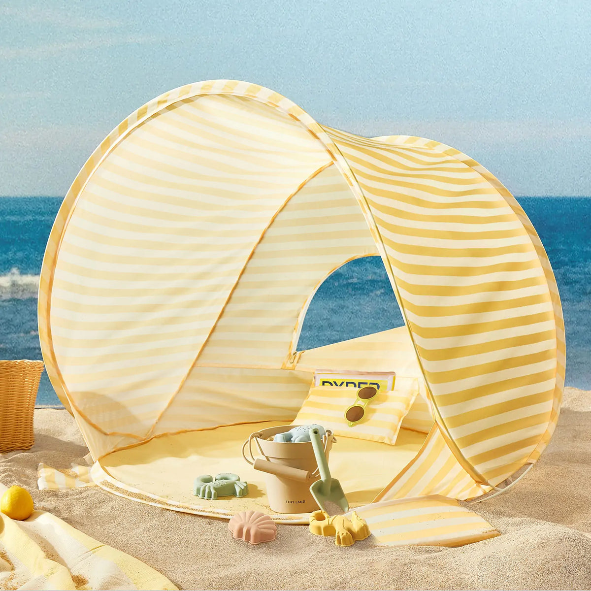 The Tiny Land® One-Stop Beach Toy & Tent Set in pastel yellow stripes sits on the sand, complete with a pillow and sand play set—bucket, shovel, and molds—by the sea under a blue sky.