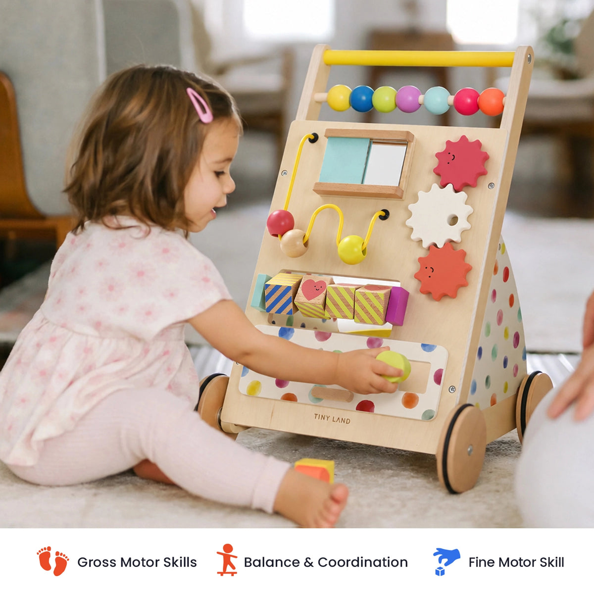 A toddler sits on the floor playing with a Tiny Land® Premium Natural Wooden Activity Walker featuring a rainbow design, bead maze, rotating gears, and shape-sorting blocks.