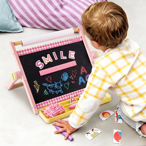 A young child in a yellow checkered shirt draws on a small chalkboard—with SMILE spelled out and colorful doodles—amidst cards and art supplies scattered across the floor. This cozy kids’ art station features the Tiny Land® Double-Sided Magnetic Tabletop Easel by Tiny Land, offering an adjustable easel with a magnetic whiteboard to inspire creativity at every turn.|Pink