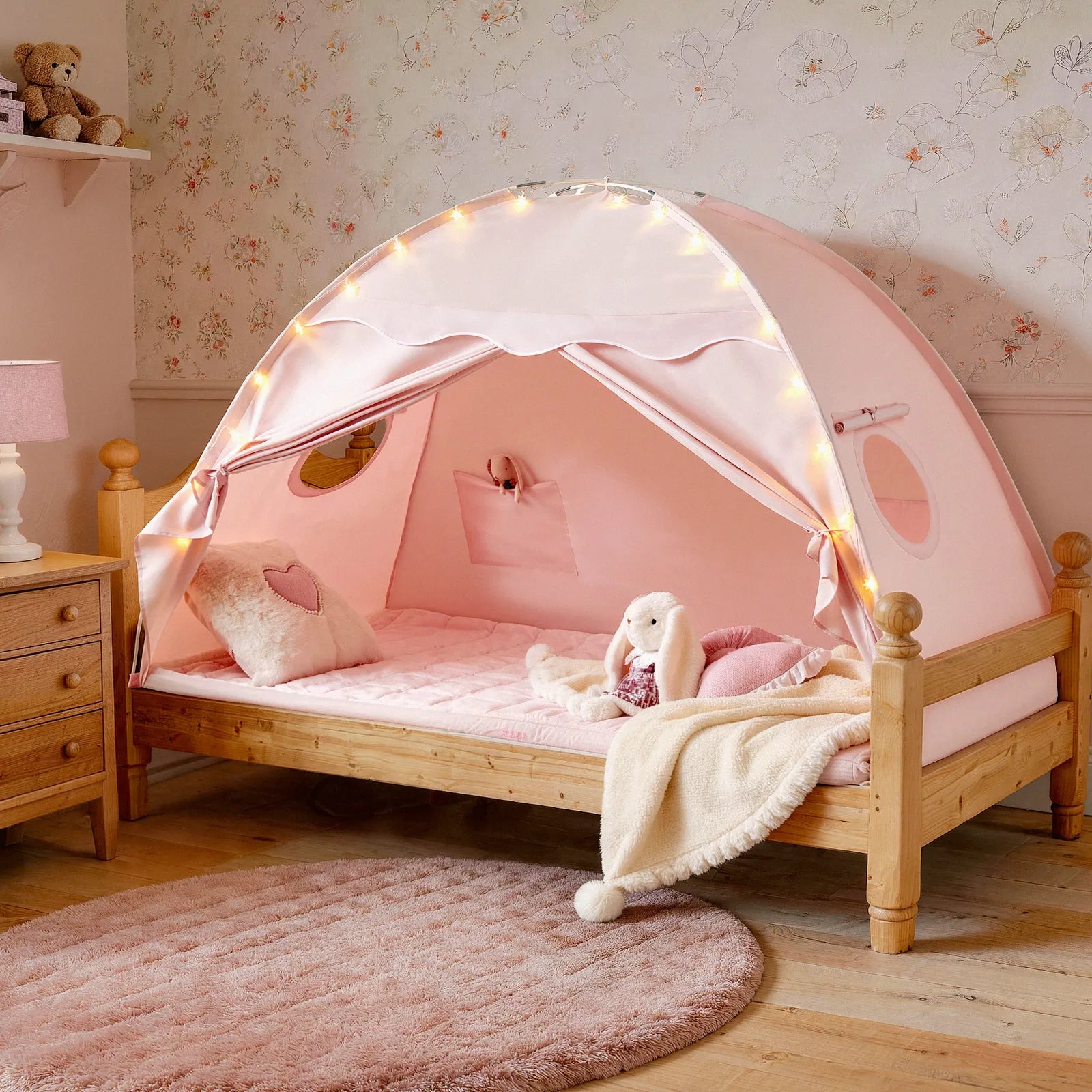 The Tiny Land® Kids Bed Tent by Tiny Land creates a playful hideaway with its pink canopy, string lights, heart pillow, and plush bunny. Its wooden frame sits by floral wallpaper, a pink rug, and a wooden nightstand with lamp.|Pink