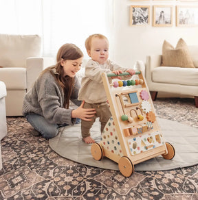 A woman smiles as she helps a toddler stand and push the Tiny Land Complete Development Playset — Baby Walker + Activity Cube across a rug in a cozy, sunlit living room, encouraging the child's fine motor skills through play.