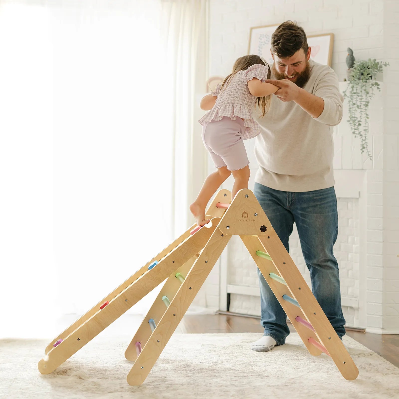 A bearded man helps a young girl balance atop a Tiny Land® Pikler Triangle 7-in-1 Montessori Climbing Set with colorful steps in a bright, cozy living room.