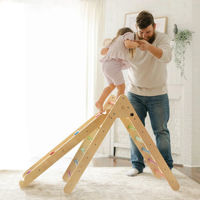 A bearded man helps a young girl balance atop a Tiny Land® Pikler Triangle 7-in-1 Montessori Climbing Set with colorful steps in a bright, cozy living room.