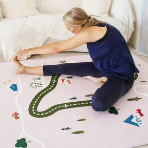 A woman in a navy tank top and leggings stretches on the Tiny Land® Double-Sided Baby Playmat Forest Track Wonder by Tiny Land, featuring road and house patterns, in a bright, cozy room.