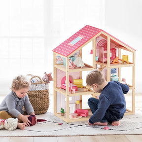 Two young children play on a rug with the Tiny Land® Sweetwood Love Dollhouse without Dolls by Tiny Land. A woven basket of stuffed animals sits nearby, while soft natural light brightens the cozy room.