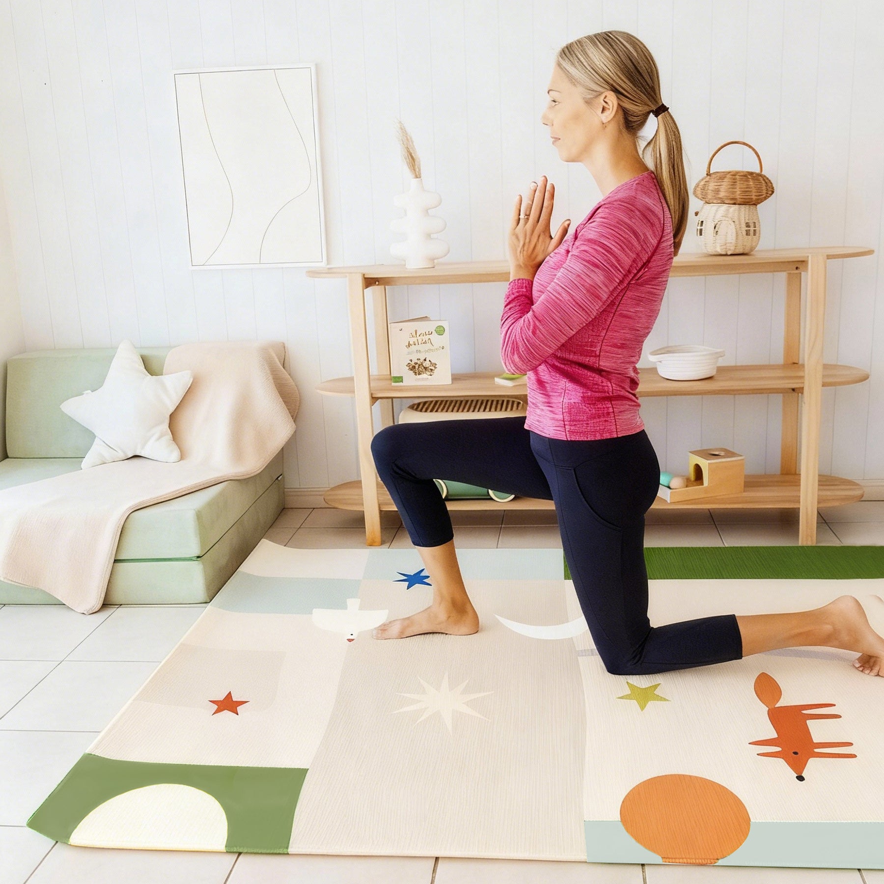 A woman in a pink top and black leggings practices yoga on the Tiny Land® Double-Sided Baby Playmat Forest Track Wonder by Tiny Land, which features playful animal and star designs. She kneels in prayer position, facing a wooden shelf and green sofa.