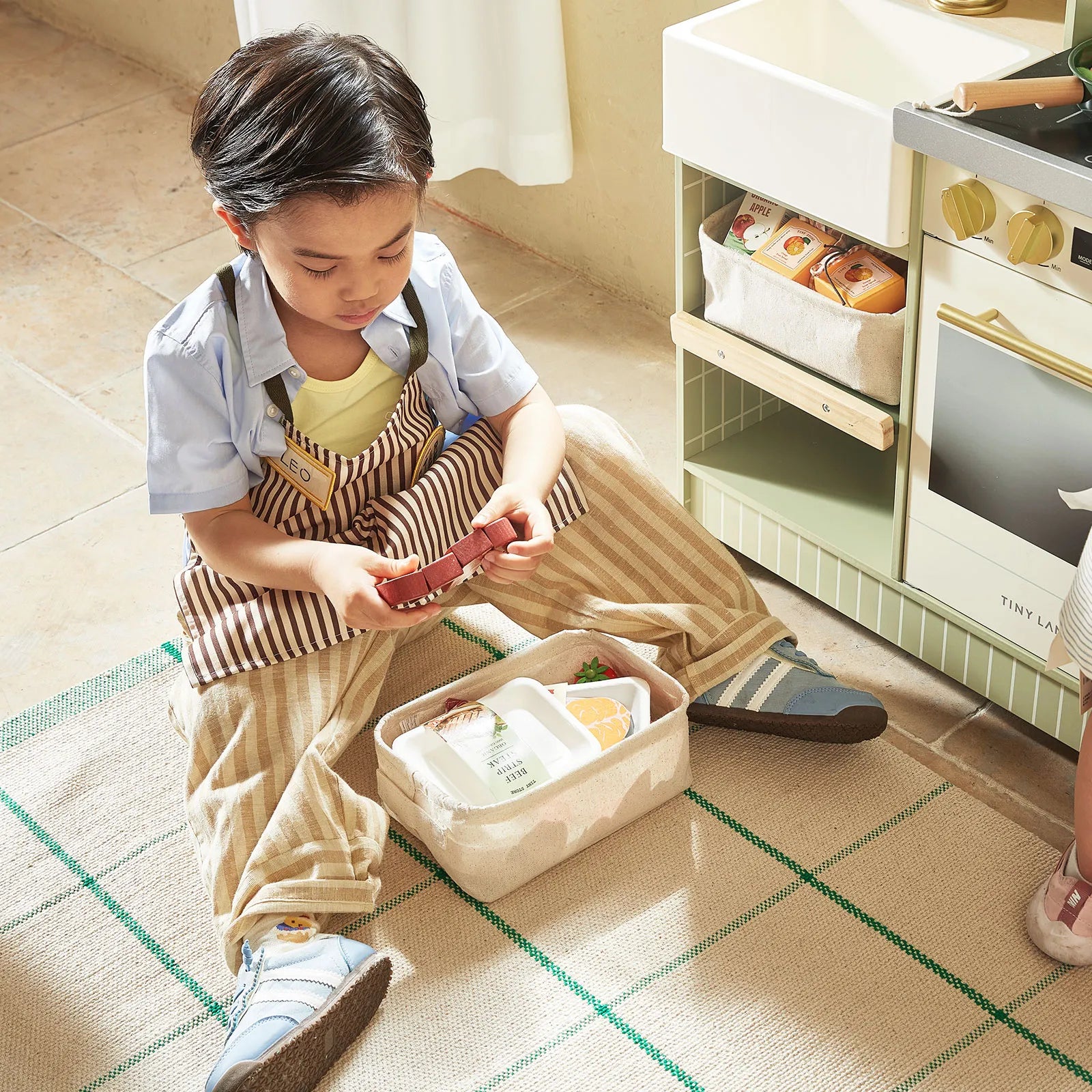 A young child sits on a rug, enjoying pretend play with toy food from a lunchbox in front of the Tiny Land® BistroSwitch Play Kitchen. Sunlight streams into the cozy room.|Green