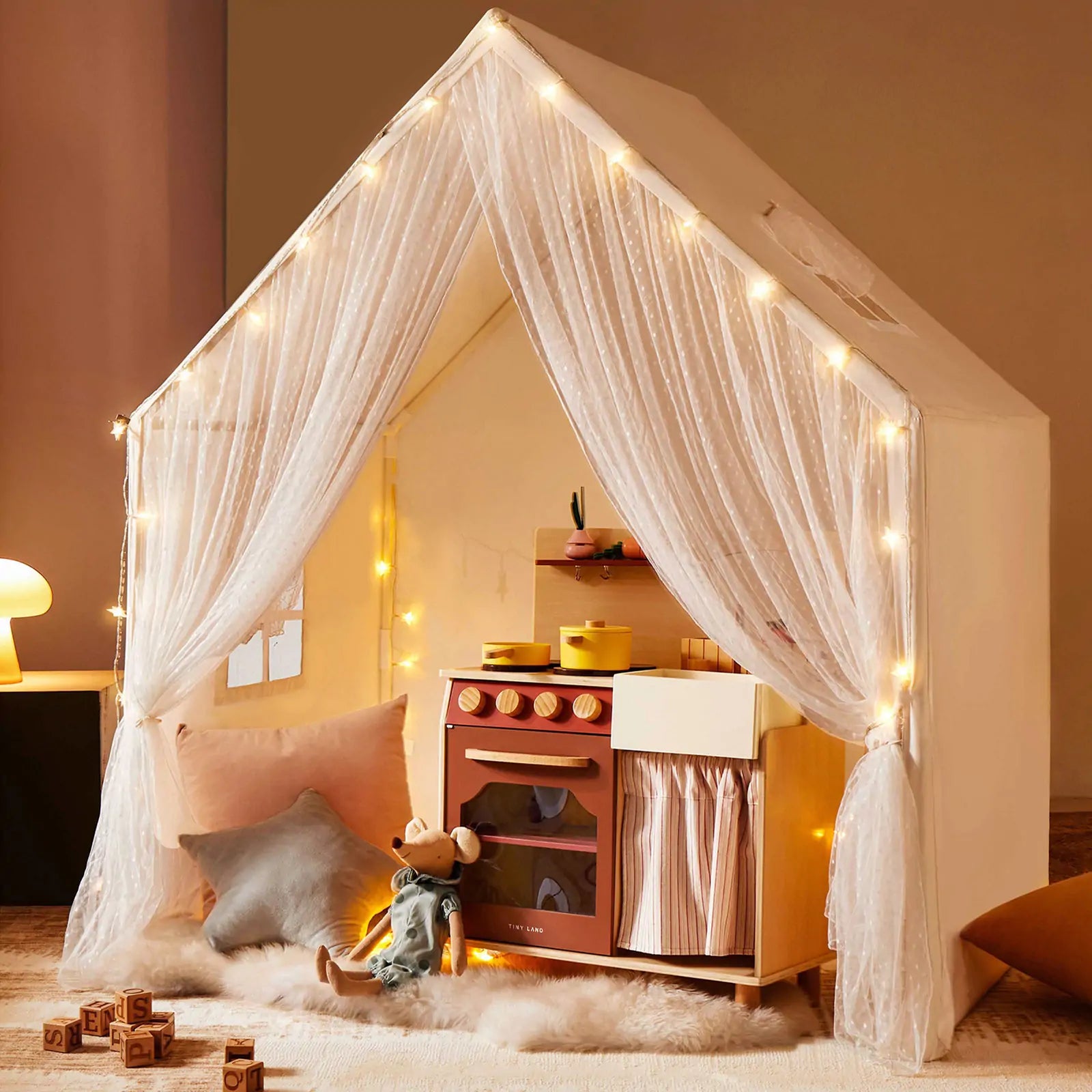 The Tiny Land® Large Space Play House with Star Lights by Tiny Land is a cozy kids’ playhouse featuring string lights, sheer curtains, a toy kitchen, plush cushions, soft rug, wooden blocks, and a stuffed mouse for inviting play.