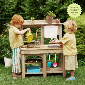 Two young children play at the Tiny Land® Mud Kitchen - DuoPlay Outdoor Play Kitchen for Kids by Tiny Land, pouring water and using toy utensils in a garden setting. Plants and kitchen items decorate the engaging sensory play setup, with a Yardzen Design Award Winner badge displayed in the top right corner.