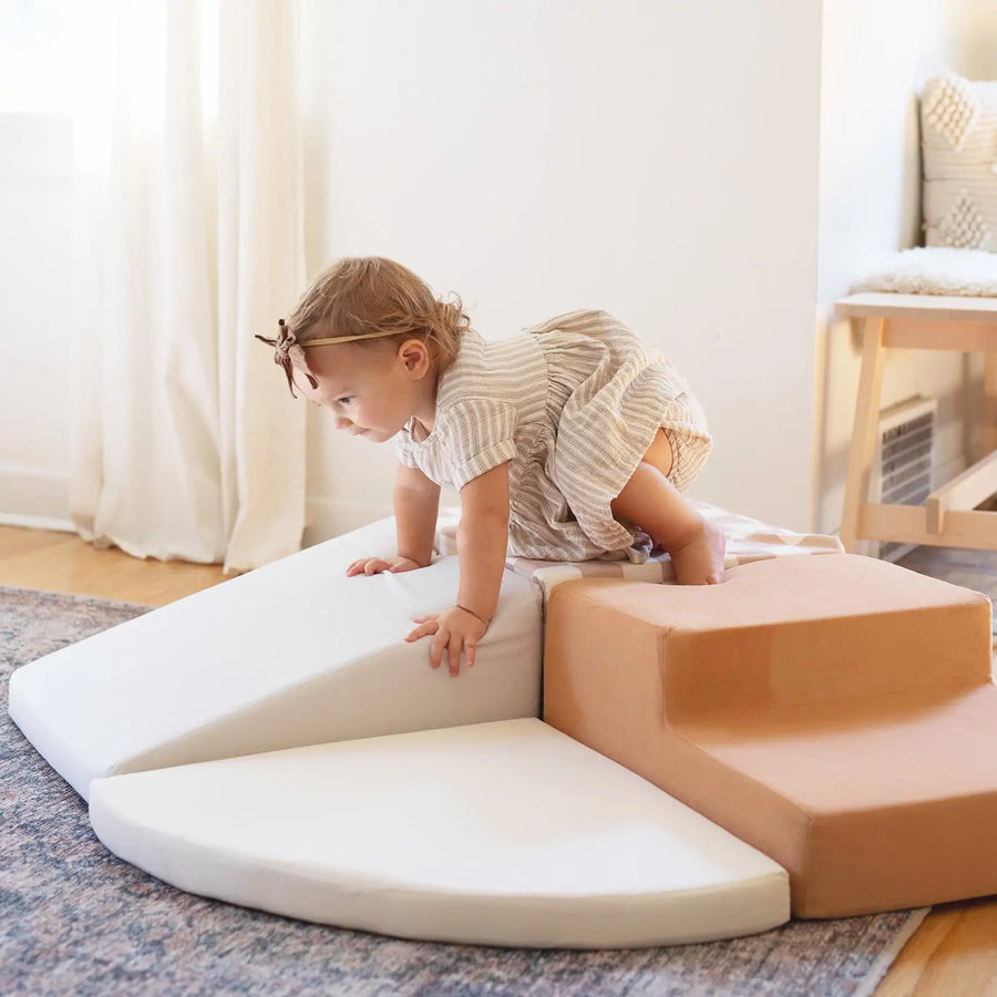A toddler in a light dress and bow climbs on Tiny Land® PlayNest Climbing Blocks by Tiny Land in a cozy, sunlit room with a rug and wooden floor, encouraging gross motor development.