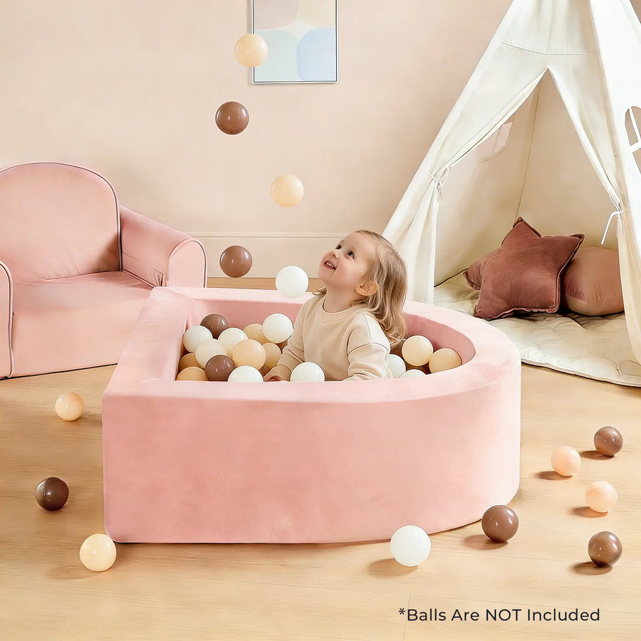 A joyful child plays in the Tiny Land® PlayNest Soft Foam Ball Pit – Balls Not Included, surrounded by brown, white, and beige balls they toss into the air. The cozy scene features a pink chair and a white play tent with pillows in the background, perfect for sensory play. Text reads: Balls Are NOT Included.|Pink