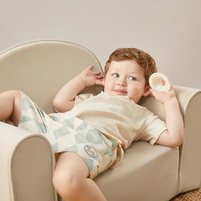 A young child with light brown hair lounges on the Tiny Land® GentleSnug Kid Chair - Beige White by Tiny Land, wearing a light shirt and patterned shorts, playfully holding a round object near their ear.