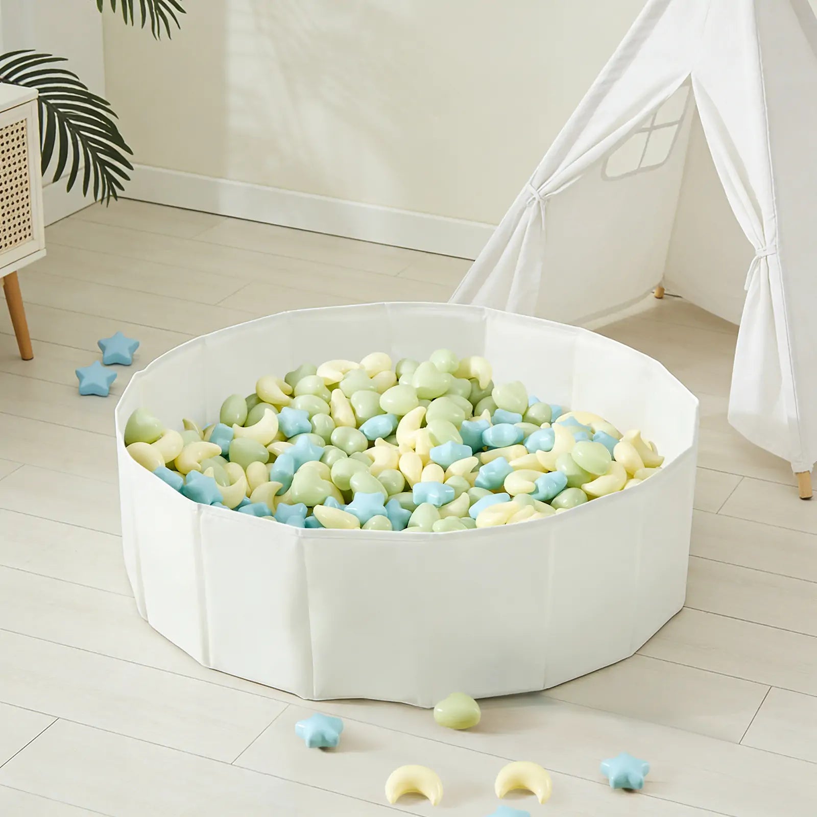 A white playpen filled with Tiny Land® Fun-Shapes Ocean Ball - 200 pcs in pastel green, yellow, and blue and star-shaped toys sits on a light wooden floor near a white teepee tent and a plant. Some Tiny Land balls are scattered outside.