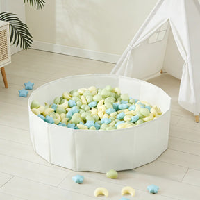 A white playpen filled with Tiny Land® Fun-Shapes Ocean Ball - 200 pcs in pastel green, yellow, and blue and star-shaped toys sits on a light wooden floor near a white teepee tent and a plant. Some Tiny Land balls are scattered outside.