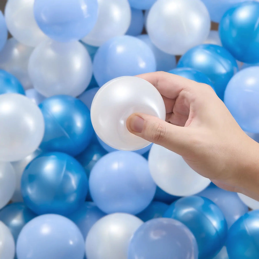 A hand holds a shiny white Tiny Land® Ocean Ball above a pile of blue, white, and light blue non-toxic balls from the Tiny Land® Ocean Ball - 200 pcs with Varied Colors set, perfect for fun ball pits or play areas.|Blue