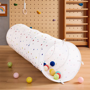 The Tiny Land® Adventure Play Tunnel - Colorful Polka Dots by Tiny Land sits on a wooden floor filled with vibrant balls, while a wooden climbing wall and gym rings make the perfect indoor play area.|white