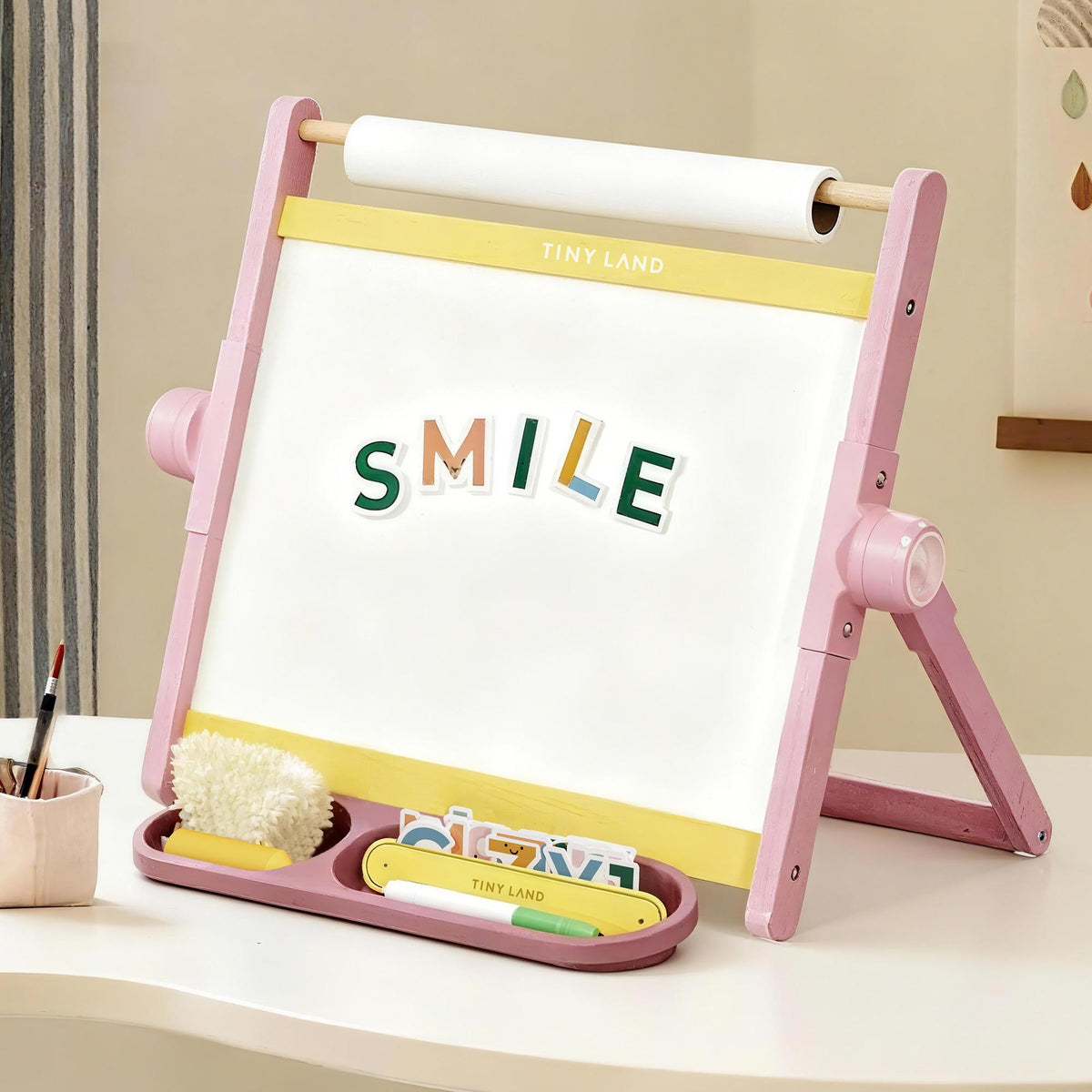 The Tiny Land® Double-Sided Magnetic Tabletop Easel - Pink by Tiny Land features a pink and yellow design with "SMILE" spelled in magnetic letters, art supplies and a fluffy duster in the tray, plus a top-mounted paper roll for creative play.