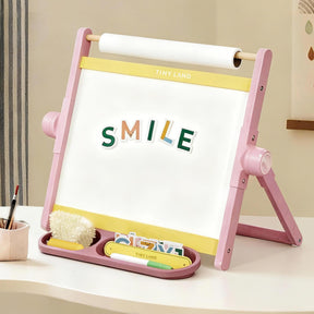 The Tiny Land® Double-Sided Magnetic Tabletop Easel - Pink by Tiny Land features a pink and yellow design with "SMILE" spelled in magnetic letters, art supplies and a fluffy duster in the tray, plus a top-mounted paper roll for creative play.