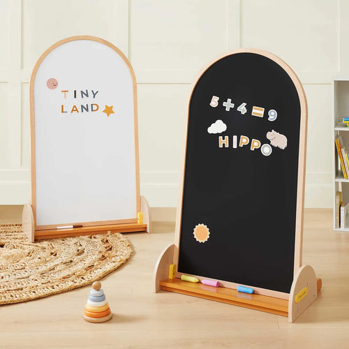 The Tiny Land® Double-Sided Arched Magnet Chalkboard from Target stands on a wooden floor; one side is a whiteboard with “TINY LAND” and a star, the other features colorful magnetic numbers, letters, and magnets. Chalk pieces and toys are nearby.