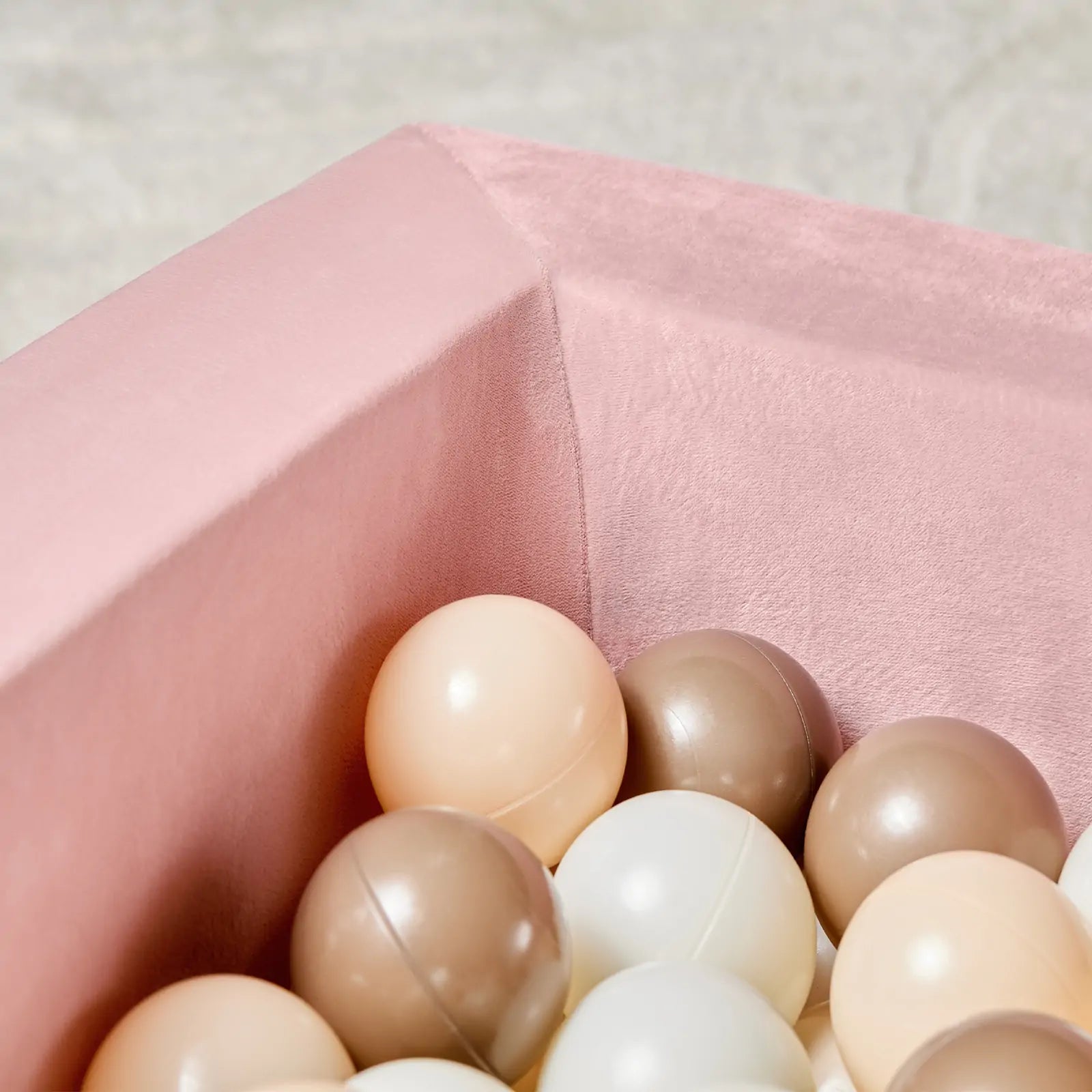 A close-up of the Tiny Land® PlayNest Ball Pit by Tiny Land, velvet-lined in pink and filled with 200 pastel ocean balls in cream, beige, and brown—perfect for creating a cozy toddler play space against a soft, neutral backdrop.|Pink