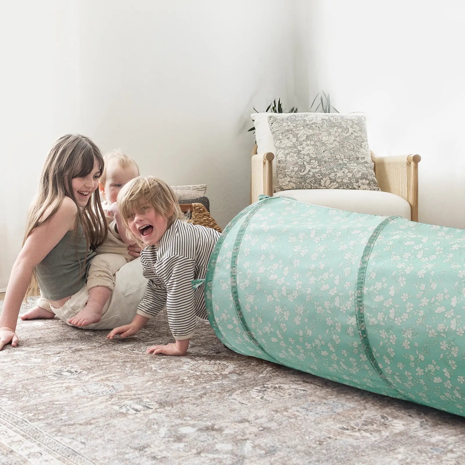 Three young children play on a patterned rug beside the Tiny Land® Adventure Play Tunnel - Green by Tiny Land. Two kids sit and smile while one crawls out of the tunnel, with a beige cushioned chair in the background adding to their fun.|green