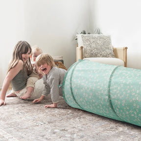 Three young children play on a patterned rug beside the Tiny Land® Adventure Play Tunnel - Green by Tiny Land. Two kids sit and smile while one crawls out of the tunnel, with a beige cushioned chair in the background adding to their fun.|green