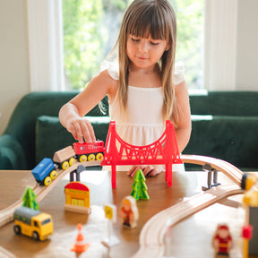A young girl plays with the Tiny Land® Wooden Train Set for Children (39 pcs) on a table. The eco-friendly set by Tiny Land includes a red bridge, trees, figures, and vehicles. Sunlight streams through the windows behind her.
