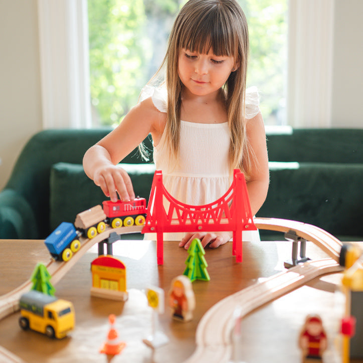 A young girl plays with the Tiny Land® Wooden Train Set for Children (39 pcs) on a table. The eco-friendly set by Tiny Land includes a red bridge, trees, figures, and vehicles. Sunlight streams through the windows behind her.