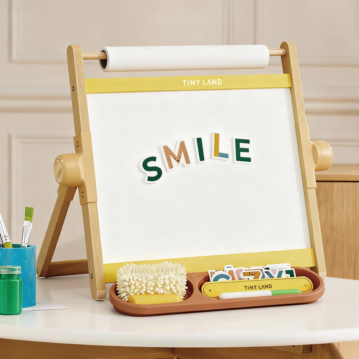 The Tiny Land® Double-Sided Magnetic Tabletop Easel - Yellow by Tiny Land displays colorful letters spelling SMILE on its whiteboard. A front tray holds a brush, letter cards, and art supplies. A beige wall and table are in the background.