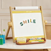 The Tiny Land® Double-Sided Magnetic Tabletop Easel - Yellow by Tiny Land displays colorful letters spelling SMILE on its whiteboard. A front tray holds a brush, letter cards, and art supplies. A beige wall and table are in the background.