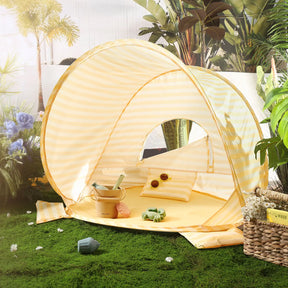 The Tiny Land® One-Stop Beach Toy & Tent Set – Pastel Yellow Stripes features a compact striped tent on artificial grass, with a pillow, sunglasses, pillow roll, and a pastel sand play set bucket surrounded by potted plants and flowers.