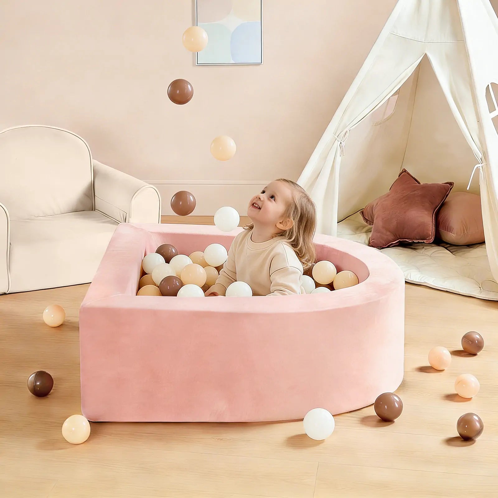 A young child smiles and looks up while playing in the Tiny Land INC Early Bird Eggs-Tiny Land® PlayNest Ball Pit filled with 200 beige, white, and brown ocean balls. The room has a beige couch and a cream play tent with cushions.|pink