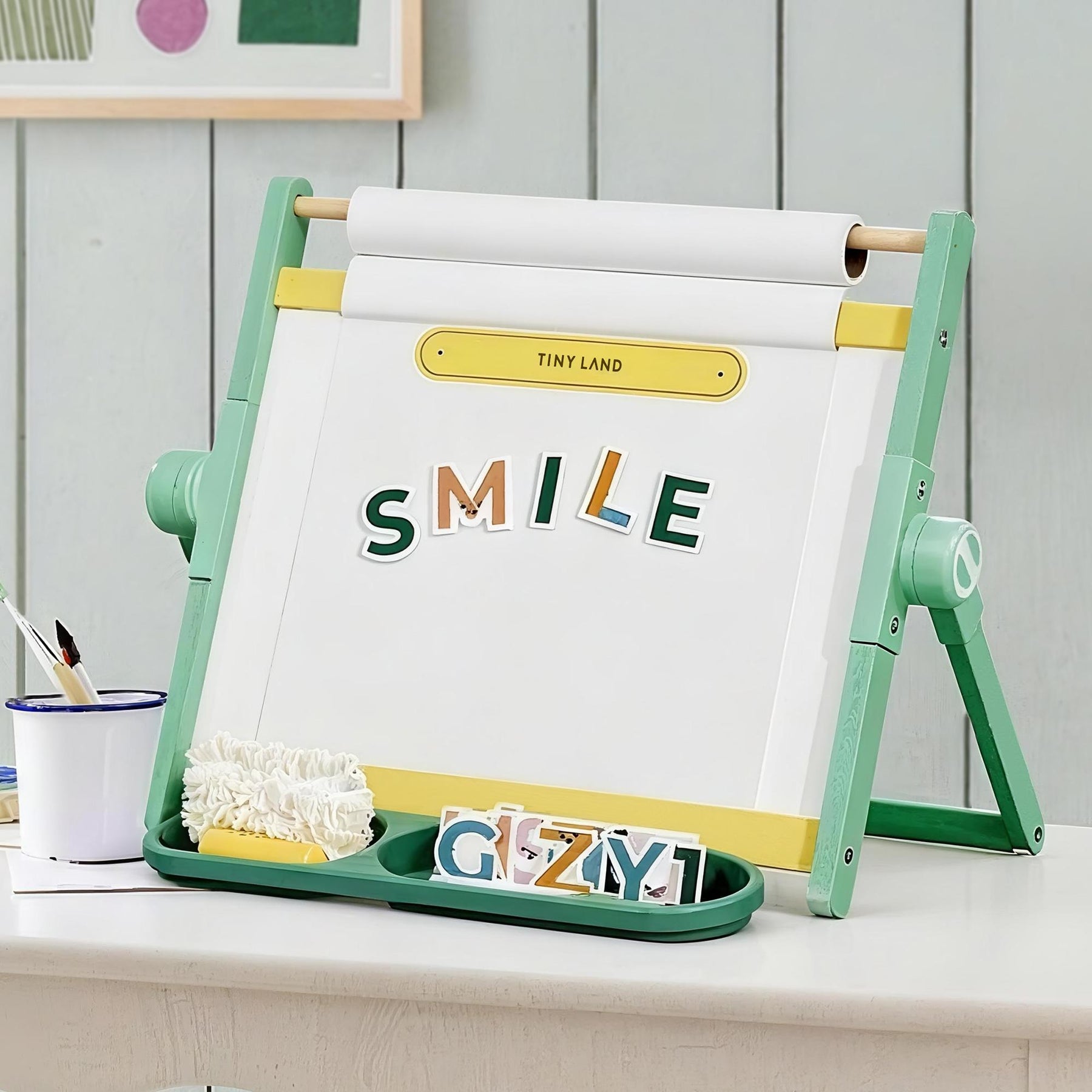 The Tiny Land® Double-Sided Magnetic Tabletop Easel - Green by Tiny Land displays "SMILE" with colorful magnetic letters on a whiteboard. A tray below holds extra letters, a white eraser, and a cup of paintbrushes for creative fun.