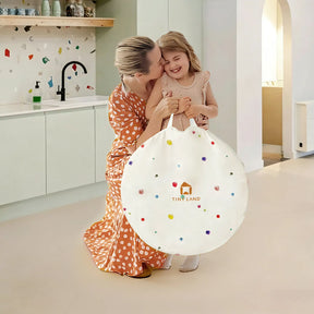 A woman in a polka dot dress hugs a smiling girl holding the Tiny Land® Adventure Play Tunnel—Colorful Polka Dots by Tiny Land, perfect for playtime storage, in a bright, modern kitchen.