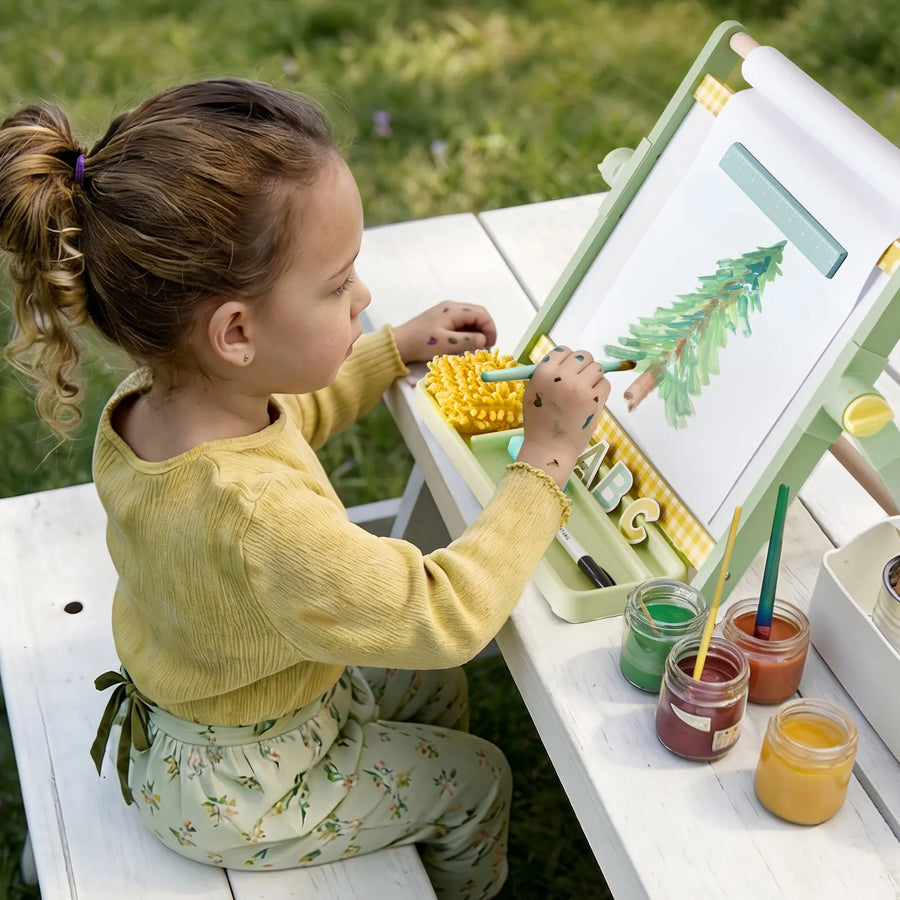 A young child sits at a white table outdoors, painting a green tree on the Tiny Land® Double-Sided Magnetic Tabletop Easel by Tiny Land with watercolor paints. Jars of paint and brushes are arranged beside her at the kids art station, with grass visible in the background.|Green