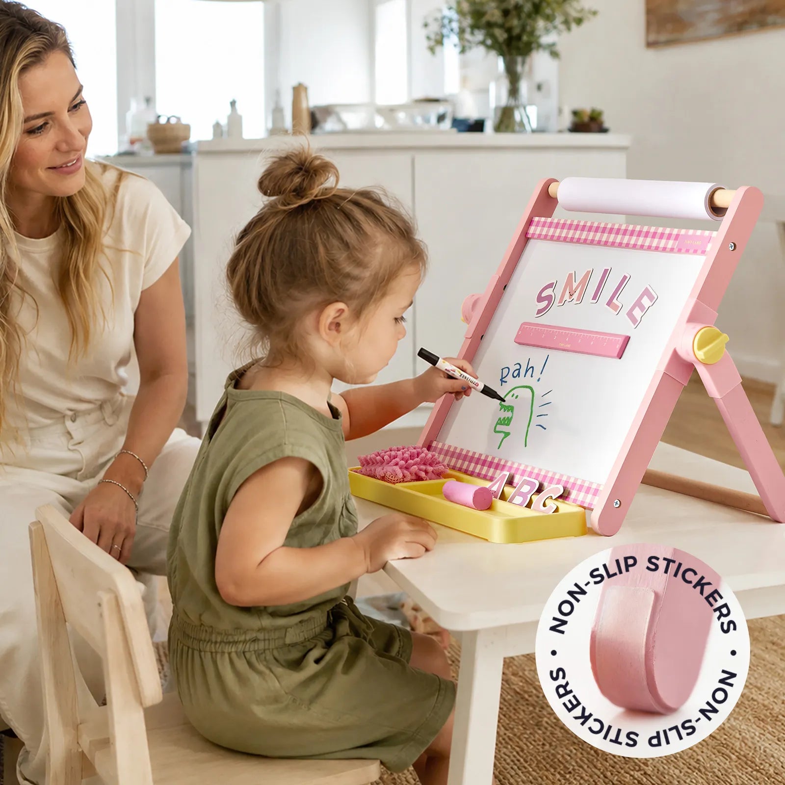 A young girl draws “SMILE” and a green character on the Tiny Land® Double-Sided Magnetic Tabletop Easel by Tiny Land, which features a pink frame. A woman sits nearby, smiling, while non-slip stickers for the kids art station are shown in the corner of the image.|Pink