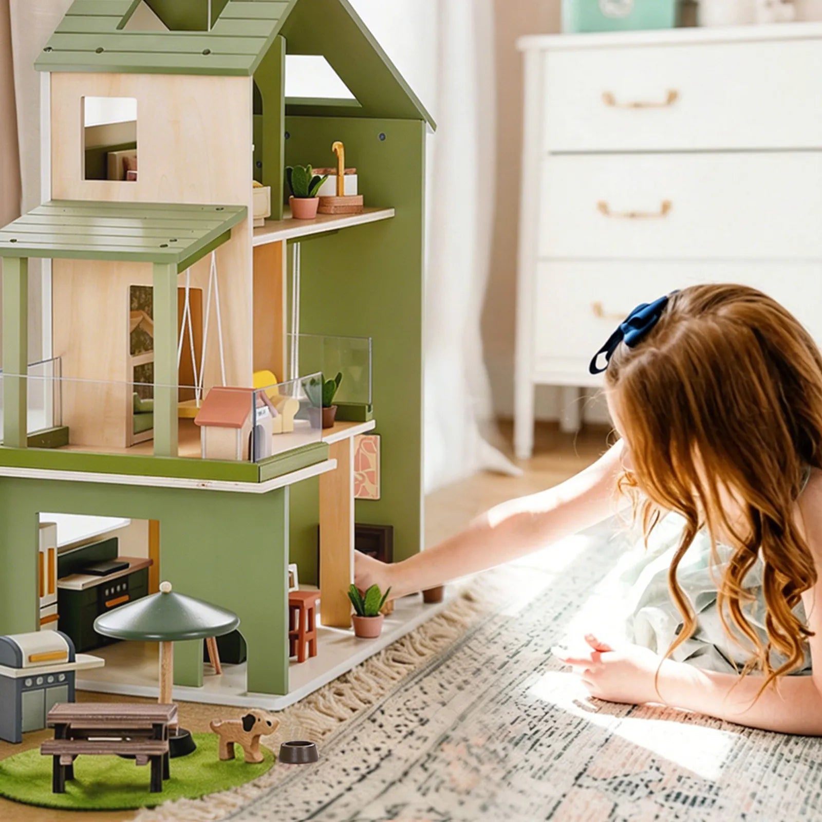 A young girl with long hair and a blue bow arranges miniature furniture and plants in the Tiny Land® Sweetwood Cottage Dollhouse on a patterned rug, with a white dresser in the cozy, bright room.|Green