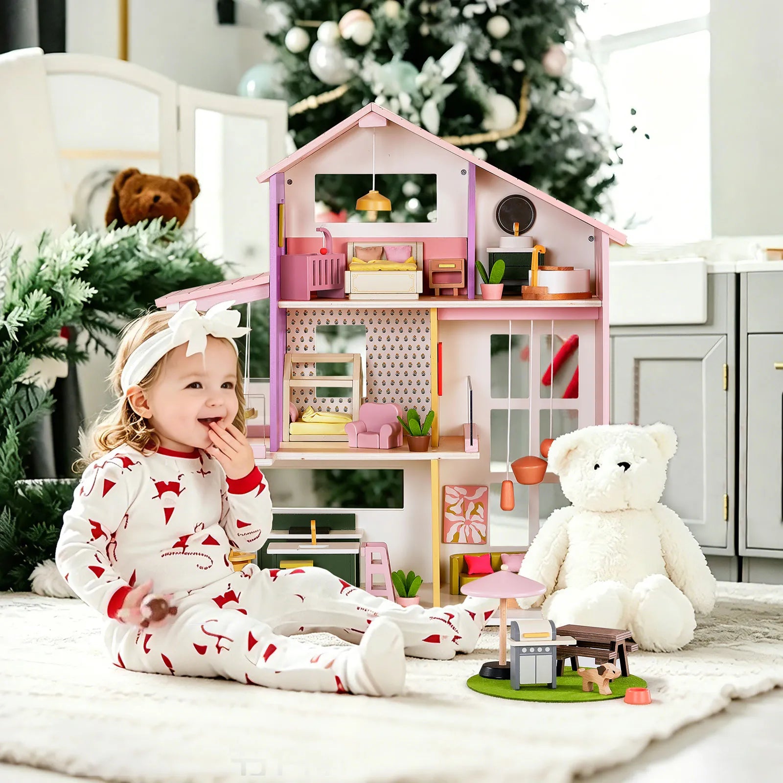 A young child in festive pajamas sits on a rug beside a large white teddy bear, smiling in front of the Tiny Land® Sweetwood Cottage Dollhouse. A decorated Christmas tree and toys add to the cozy, well-lit room.|Pink