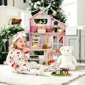 A young child in festive pajamas sits on a rug beside a large white teddy bear, smiling in front of the Tiny Land® Sweetwood Cottage Dollhouse. A decorated Christmas tree and toys add to the cozy, well-lit room.|Pink