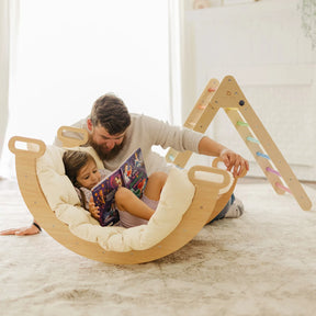A bearded man and a young girl sit in a cushioned rocker, reading together. Nearby, the Tiny Land® Pikler Triangle 7-in-1 Montessori Climbing Set by Tiny Land rests on a soft rug—perfect for an indoor playground in this cozy, sunlit room.