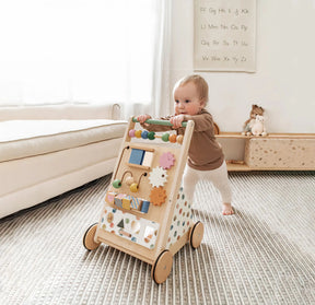 A baby pushes the Tiny Land® Versatile Natural Wooden Baby Walker by Tiny Land across a striped rug in a cozy, softly lit room decorated with toys and a letter chart on the wall.